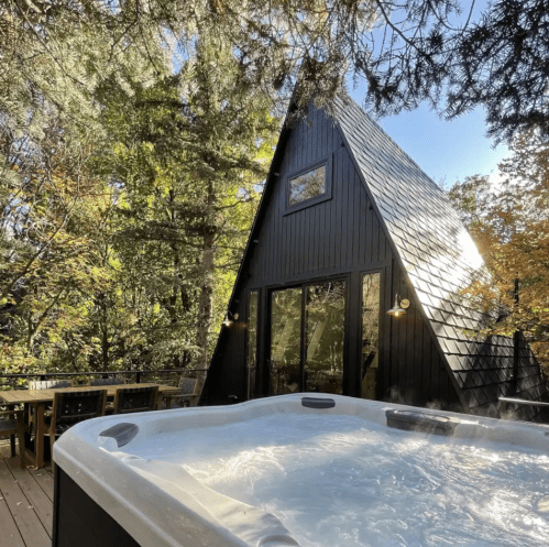 A modern A-frame cabin surrounded by trees, featuring a hot tub on a wooden deck.